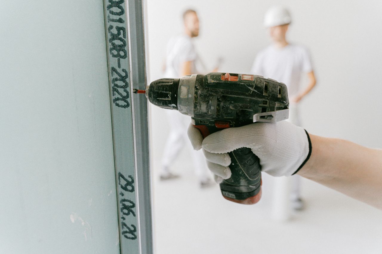 Home A construction worker uses a drill on drywall with blurred workers in the background.